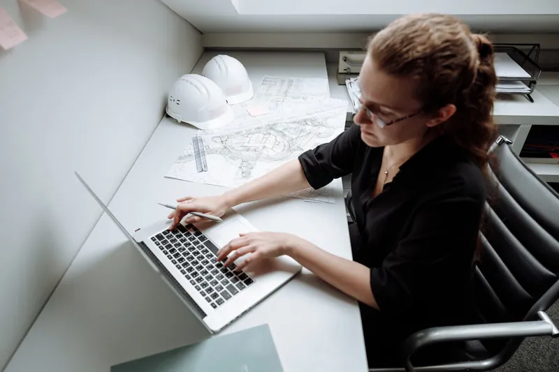 An engineer working at a laptop with drawings and a hard hat on the desk